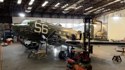 The Douglas C-47 Skytrain a dark green design with white printing on its side and front. It is in a warehouse during restoration work.