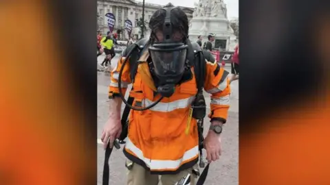 Gilly Photography A man dressed in firefighter kit including mask and breathing apparatus standing in front of Buckingham Palace with marathon runners in the background