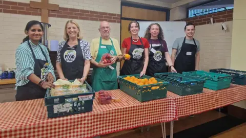 BBC/Naj Modak FoodCycle volunteers pose behind tables with crates of oranges, apples, and other produce while wearing aprons in a community hall, ready to prepare food for a community meal. Ian White is stood with them with glasses, a yellow t-shirt and green apron.