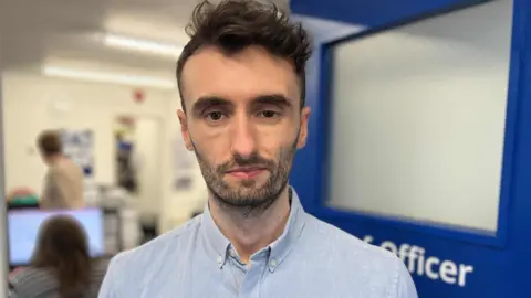 Fraser Gleave looking at the camera, is wearing a blue shirt and has an office in the background.