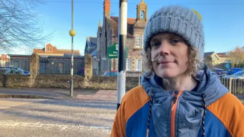 A woman with blonde hair popping out from under a beanie hat, wearing a blue and orange weather proof jacket stands beside a faded zebra crossing.