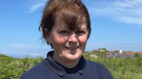 A wide shot of Angela Salmon. She has short brown hair and is wearing a navy polo shirt and jumper.