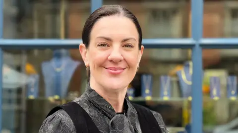 Daisy Coppin is smiling for the camera. She has dark hair which is neatly pulled back into a ponytail. She is wearing a black and white patterned blouse with a black waistcoat over the top. 