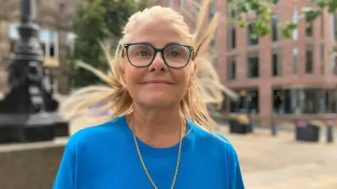 Steve Jones/BBC Jill Brodie - a woman with blonde hair and glasses. She is wearing a blue top and is smiling at the camera.
