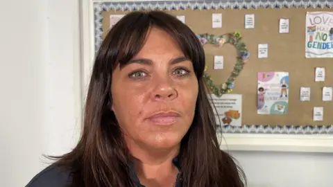 Danielle Chadwick has shoulder length brown hair and is looking serious. She is stood in front of a notice board with a love heart pinned up on it.