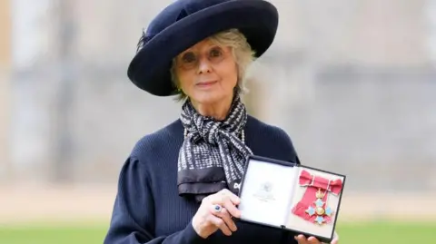 Getty Images Diana Parkes in a dark blue top and matching hat holding her CBE medal with its red ribbon