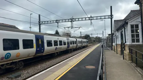 Jessica Bayley/BBC A Northern rail train on the left side of the track, picture has been taken from the station platform, can see the entrance to the right hand sided of the picture.