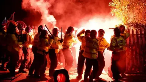 Reuters Officers in riot gear holding up batons and circular shields as red flares are thrown at them