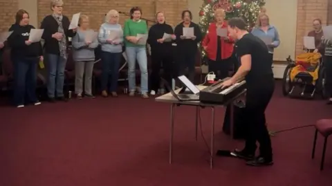 About 10 people holding song sheets and singing. A man in the middle is playing keyboards. 