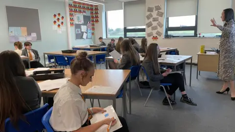 Pupils being taught in a classroom. They are sitting on blue plastic chairs at wooden desks.