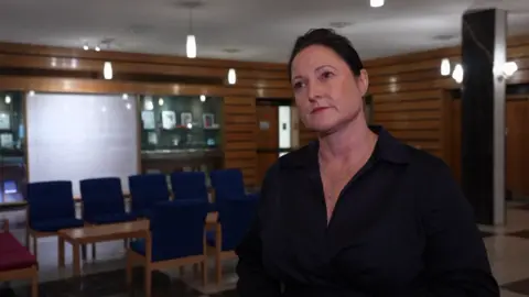 A woman in a black, open-necked blouse with tied back dark hair stands in a wood-panelled room full of blue chairs.