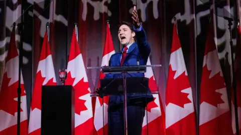 Reuters Perdana Menteri Kanada Justin Trudeau berbicara di podium pada pesta liburan kaukus federal Liberal, mengenakan setelan jas dengan sejumlah bendera Kanada di belakangnya