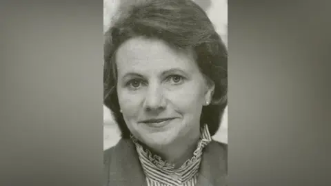 Joanna Spicer A black and white image of Joanna. A head shot showing her with dark hair and pearl earrings, a striped shirt with high collar and a dark coloured jacket.