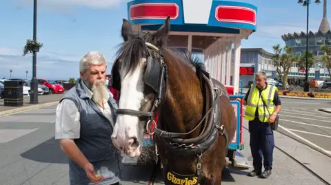Manx Scenes A driver with a long grey beard stands next to a brown horse, bearing the nameplate Charles and pulling a tram. The pair are pictured in front of the Sea Terminal.