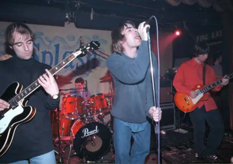 Getty Images Oasis perform live on stage in the early 1990s. Liam Gallagher sings into a microphone at the centre, flanked by bandmates playing guitar, with the drummer visible behind a red Pearl drum kit. A banner reading "Splash" hangs in the background.