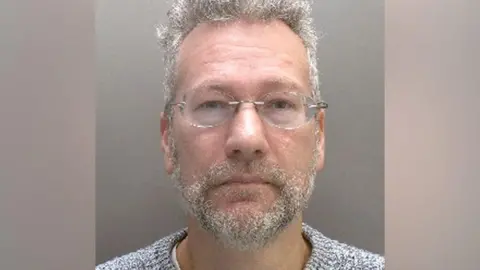 Mugshot of man with grey hair and beard and glasses