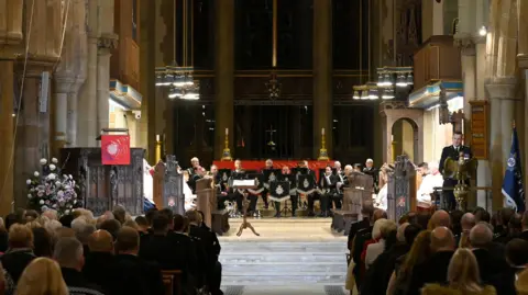 West Yorkshire Police The inside of a large cathedral with high stone arches and tall columns. In the foreground, there are rows of seated people sat in pews, facing forward. They are looking on at a group of uniformed musicians seated in a semi circle, holding brass instruments.