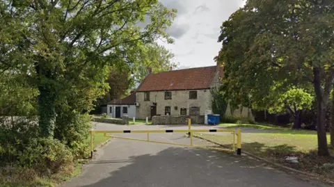 Google A general view of the Whitchurch Folk House in Bristol, seen from the nearest street. It is framed by trees and has a yellow and blue traffic barrier in front of it. The building itself is made of light brick and has a red tiled roof and black-framed windows