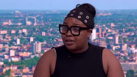 A woman in a black armless dress with a black band on her head sitting in a TV studio being interviewed. 