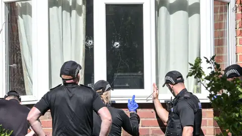 Alamy Five police officers in black uniforms and baseball style caps carry out a forensic examination. They are looking at two bullet holes in the window of a house 