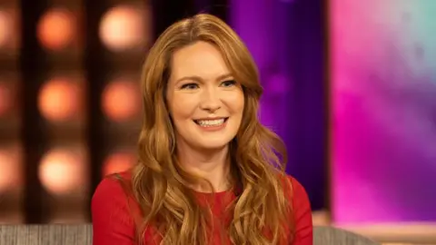 Picture of Sarah J Maas in a red top. She has ginger hair and is sat in front of a blurred background