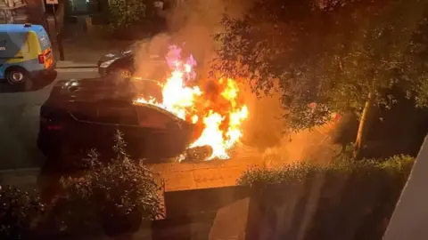 The front half of a car parked on a residential street is seen engulfed in flames. 