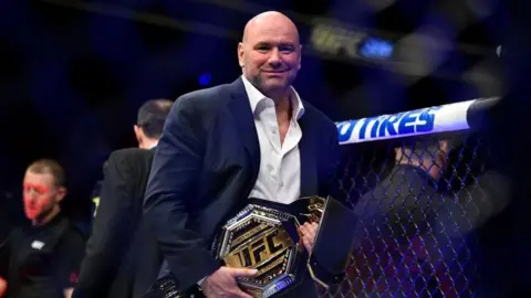UFC boss Dana White is pictured inside the octagon holding a gold UFC belt. He is wearing a blue suit with a white shirt. 