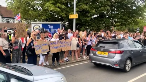 A crowd of protesters holding signs stand on the pavement outside a hotel as cars pass them on the road in front of them.