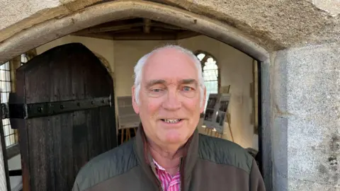 John Devine/BBC John Souter has a mainly bald head, with some white hair visible at the top which runs down the side of his face just above his ear lobes.
He has a dark fleece jacket on with a red and white striped shirt underneath, behind him is a old stone arch and doorway into a chapel.