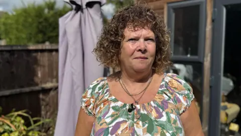 BBC Julie Parton - a woman with brown curly hair and wearing a floral dress - stands in front of a shed in a garden.