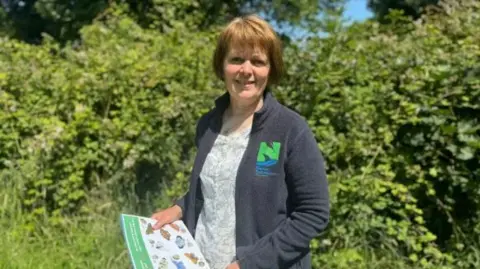 Angela Salmon smiling and wearing a grey fleece jacked with the Nature Commission logo, which is green and blue. She is stood in front of green hedges and holding a piece of paper with pictures of butterflies.