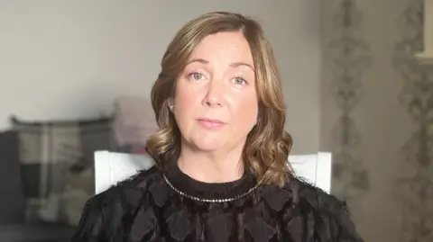 Martina Tierney sits in her living room and looks at the camera. She has shoulder-length, light coloured hair and is wearing a black shirt. She is sitting in front of a grey sofa with a patterned wallpaper.