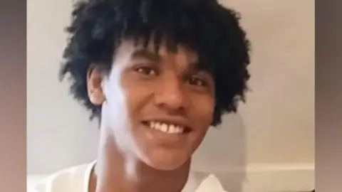 Devon Simmonds-Caines, with black mid-length curly hair and wearing white top, smiles at the camera