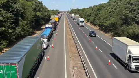 View of the M11 from a bridge over it. There are two lines of traffic, one heading away from the camera and one towards. The right lanes in both directions have been blocked off by orange traffic cones. The sky is blue and bright and there are green trees at either side of the motorway.