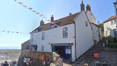 A white seaside building with a brown tiled roof houses a coffee shop and visitor centre, set beside cobblestone steps, with people walking nearby and Union Jack bunting strung overhead against a bright beach and blue sky.