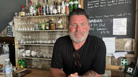 George Carden / BBC A man, Kevin Dawson, wearing a black t-shirt. He is stood in a wine bar, leaning over a counter with his arms resting on the wooden surface.