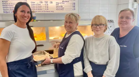 BBC Four women stand beside a chip shop fryer. Two are dressed in white tops, one in a grey jumper and one in a black t-shirt. They all wear navy tabards. There is a blue and white illuminated price list above the fryer on a white tiled wall.