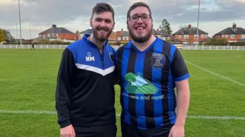 Harry McCabe and his brother Jack. They both have short brown hair and beards. Jack is wearing glasses and a blue and black football shirt while Harry is wearing a blue, white and black sports top. They are stood on a football pitch with houses in the background.