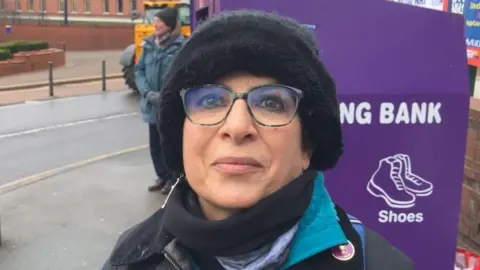 A woman with glasses wearing a black fluffy hat smiles at the camera with her mouth closed.