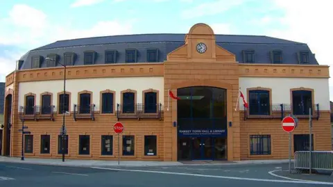 The outside of Ramsey Town Hall, which is a curved cream and caramel coloured building over three floors with a large glass-fronted entrance.