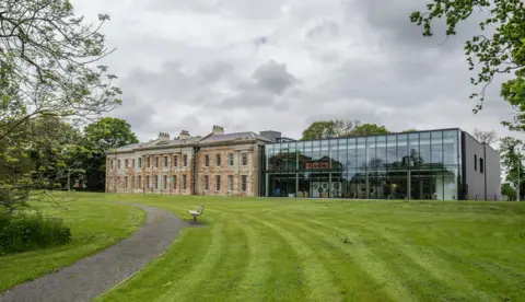 Mount Oswald Manor House. The left-hand side of the building is constructed from stone, while the right-hand side has all-glass frontage. It is surrounded by grass and trees.