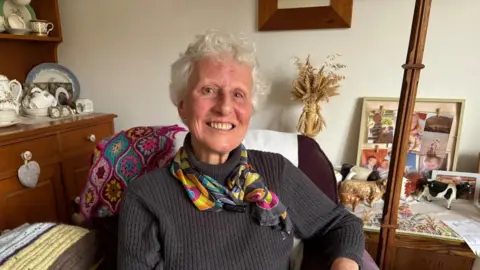 A woman with short grey hair smiles at the camera. She is wearing a dark grey jumper and and patterned scarf and sitting in a maroon arm chair. Behind her is a sideboard with a display of china on it and to her right there are some family photos and ornamental cattle.