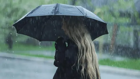 Getty Images The image shows a blonde woman under a black umbrella, wearing a black coat. In the background trees and a road can be seen blurrily.