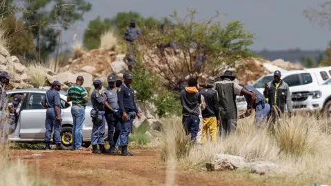 Police officers wearing blue uniform stand around at the entrance of a mine shaft where 4,500 illegal miners are trapped underground - 13 November 2024