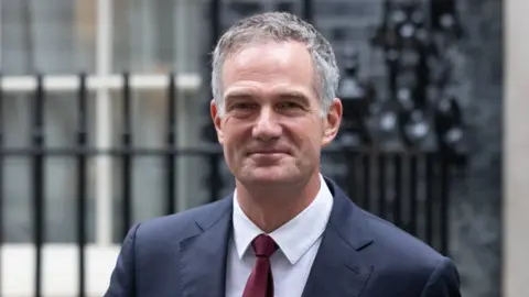 Getty Images A man with a blue jacket, tie and white shirt is looking at the camera. Railings are blurred behind him.