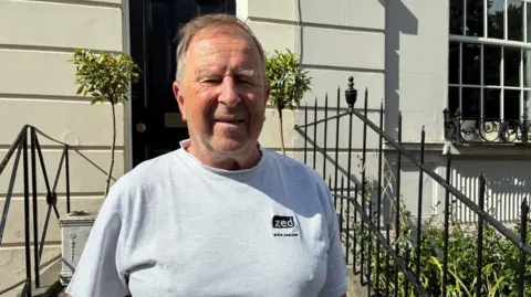 A middle aged man with short grey hair and a grey T-shirt on squints in the sun as he stands outside a lovely cream coloured townhouse, with small olive trees outside.