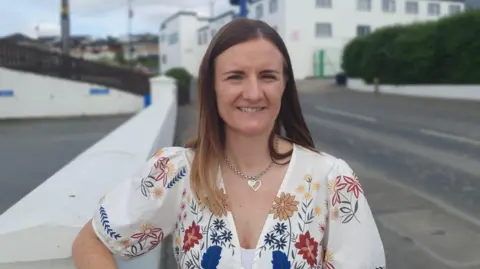 A woman in a white top, which is covered in flowers, stands with one arm on a white wall. she has long brown hair, and is wearing a silver necklace, in the background is a factory