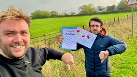 Marcus Vickers and David Reynard are standing in front of a wooden fence in a field. They have their thumbs down and Reynard is holding papers that read: "Local Plan". 