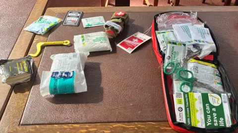 The contents of a bleed control kit, including the opened red case, with dressings and scissors inside, as well as dressings and bandages and other items on a table next to it