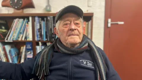 An elderly man is looking off to the side of the camera. He is wearing a navy zip up fleece, a peak cap and a striped scarf. Behind him is a book shelf and a red door. 